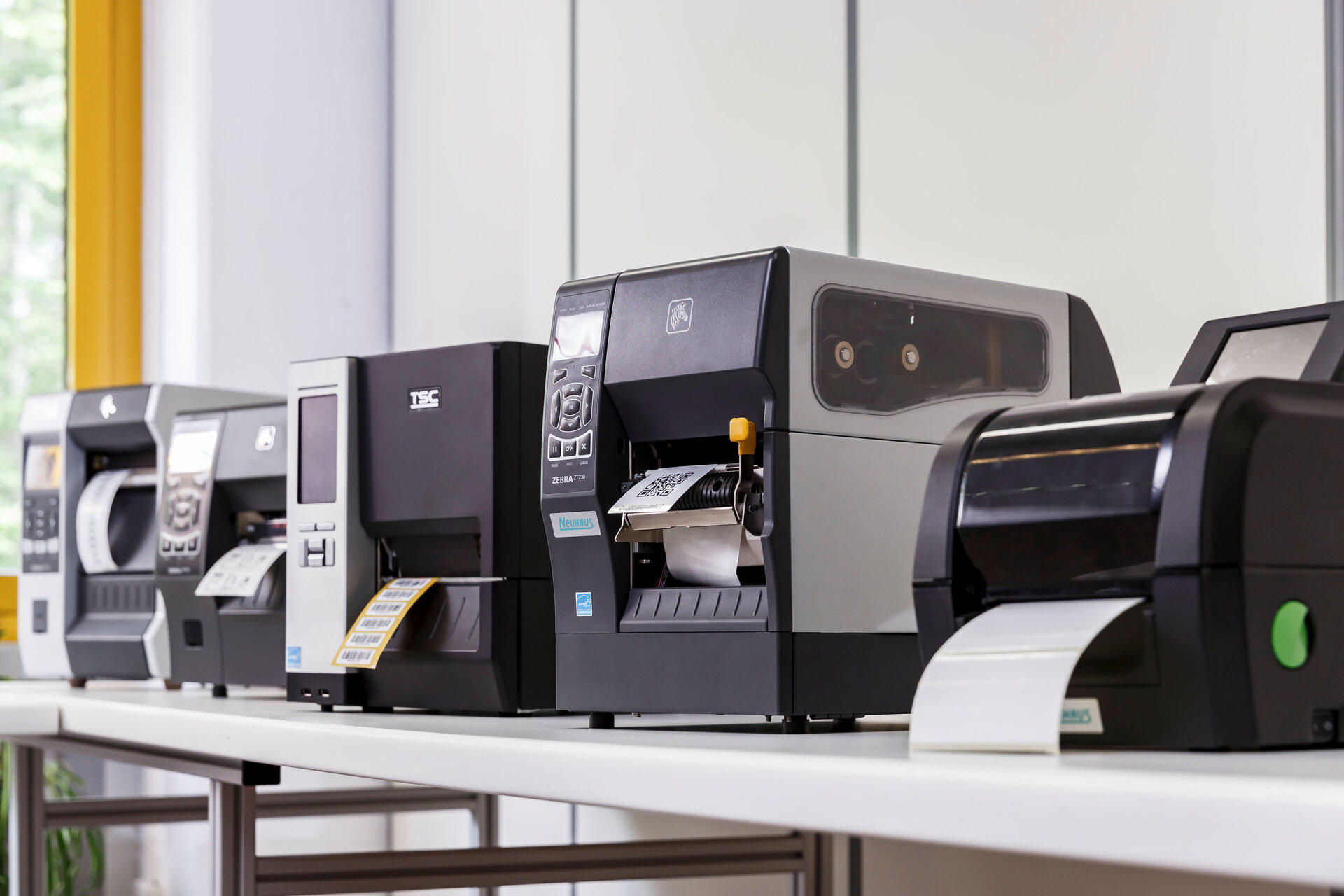 In einem hellen Raum mit großen Fenstern steht eine Reihe verschiedener moderner Etikettendrucker auf einem weißen Tisch. Jeder Drucker druckt aktiv Etiketten, und einige Etiketten kommen bereits sichtbar aus den Geräten.